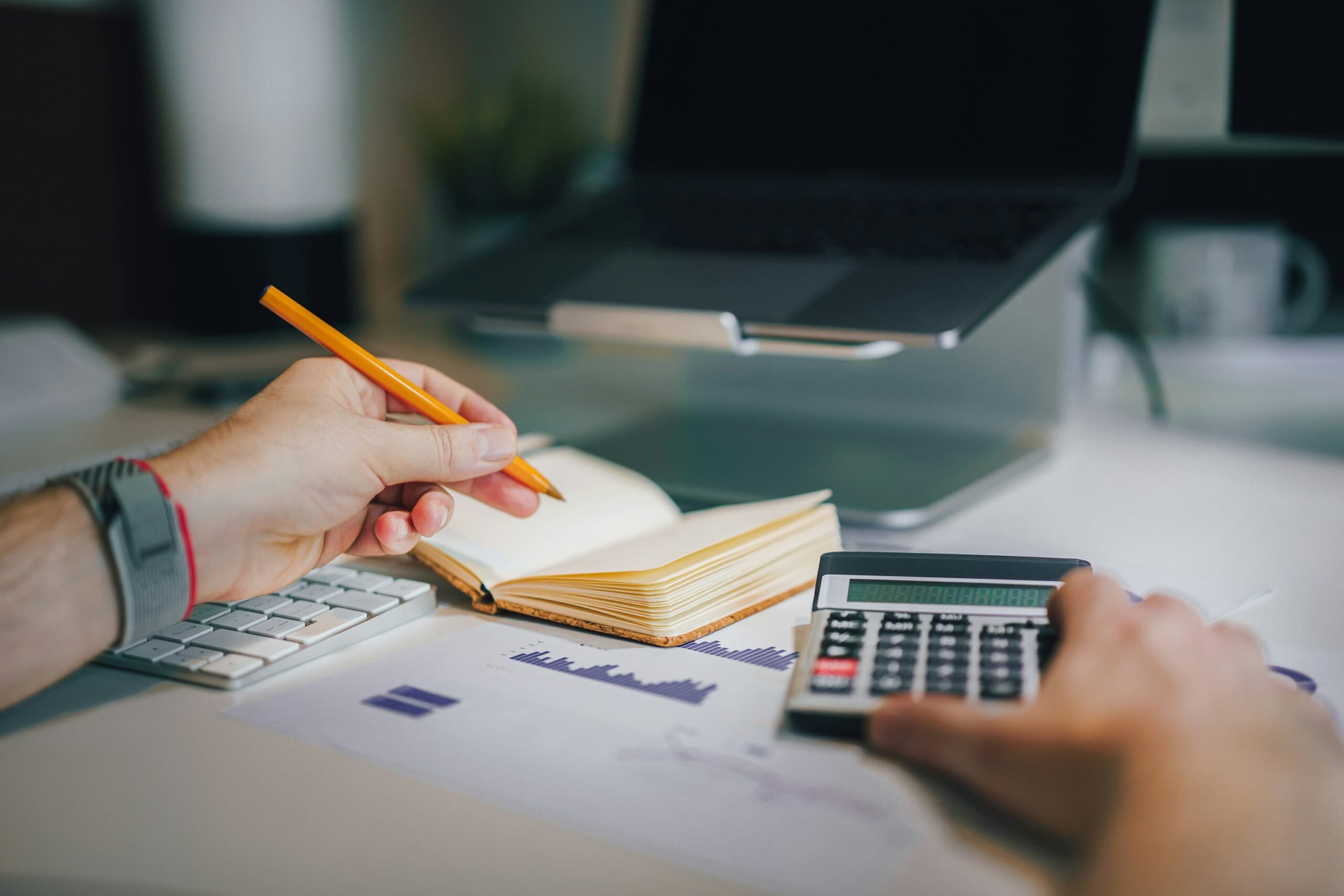 a hand holding a pencil over a note book with a calculator next to it